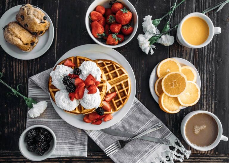 Breakfast table with waffles topped with whipped cream and berries, muffins, a bowl of strawberries, orange juice and sliced oranges