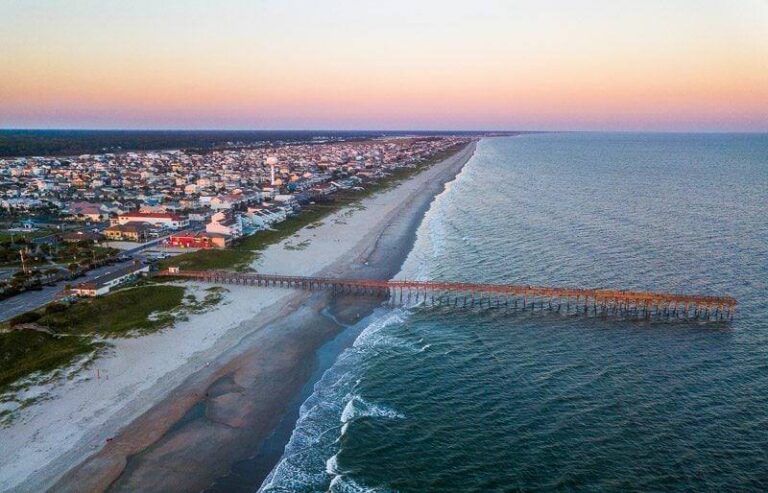 Aerial view of the Ocean Isle Beach Pier reaching into the Atlantic, with the beach and town along the shore under a pink and orange sky